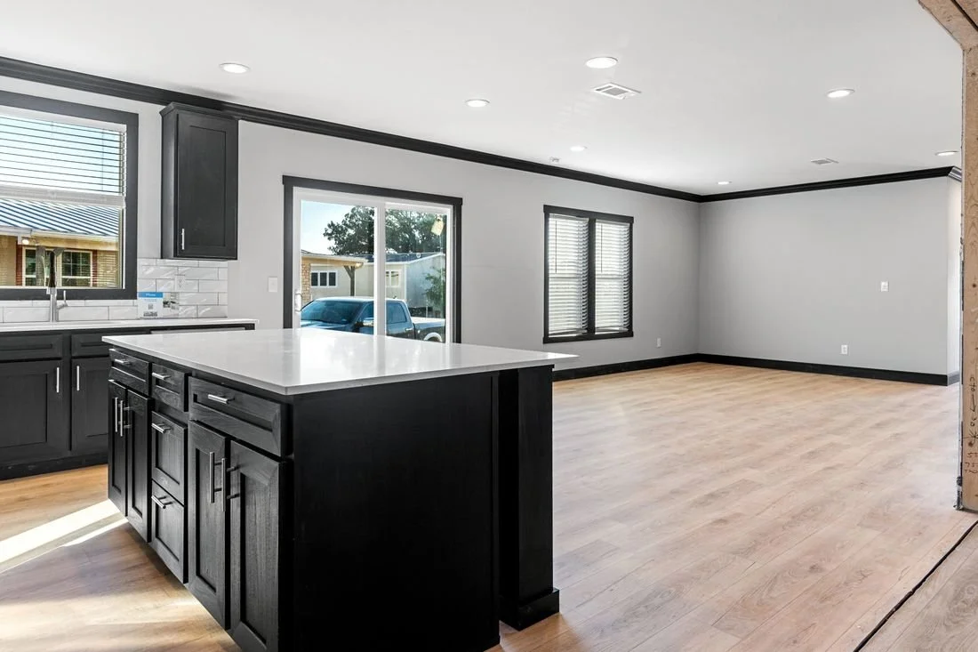 Empty modern kitchen and living area with black cabinets, white countertops, large windows, and hardwood floors.