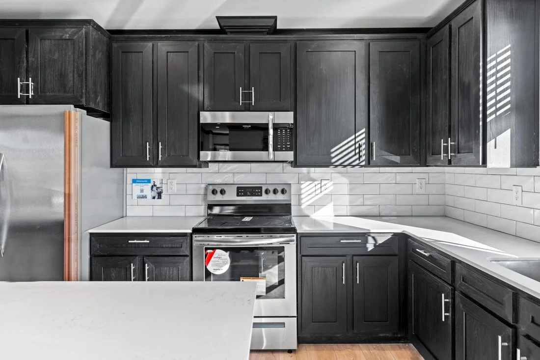 Modern kitchen with black cabinets, white countertops, stainless steel fridge and stove, microwave, and white tiled backsplash with sunlight patterns