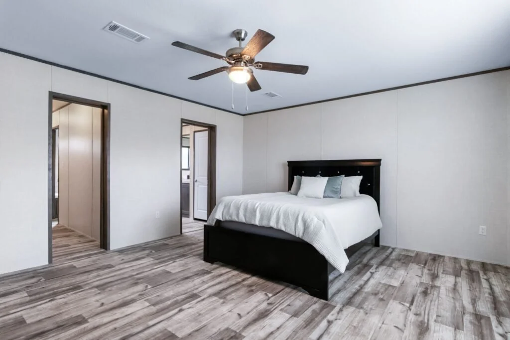 A bedroom with a large bed, dark wooden headboard, white bedding, and pillows, hardwood flooring, a ceiling fan with lights, and two open doorways leading to other rooms.