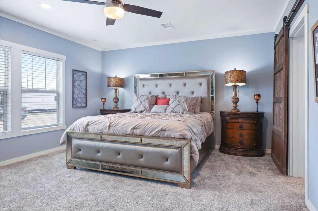 A bedroom with a large bed, tufted headboard, and matching footboard, flanked by two wooden nightstands with lamps, light blue walls, window with blinds, and beige carpet.