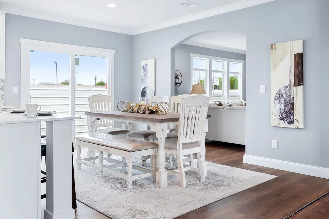 Dining room with white wooden table, six chairs, abstract art on light gray walls, hardwood floor, large window letting in natural light, and a living room area visible through an archway.