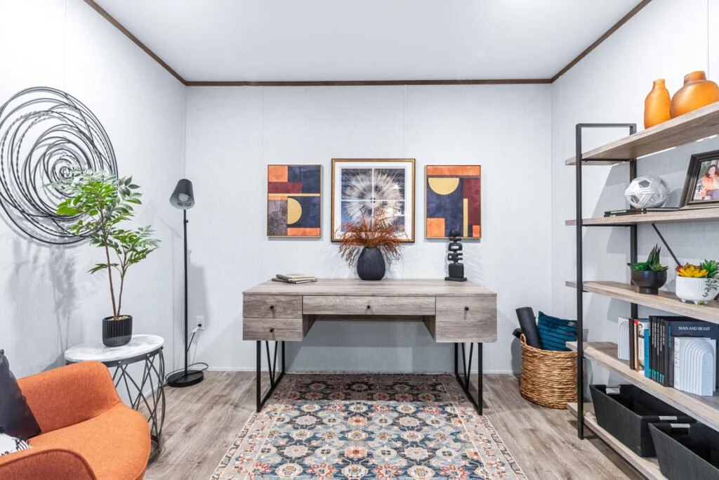 Living room with a modern decor, including a wooden desk with vases and decorative items, colorful artwork on the wall, a tall bookshelf with pottery and books, a potted plant, and an orange armchair.