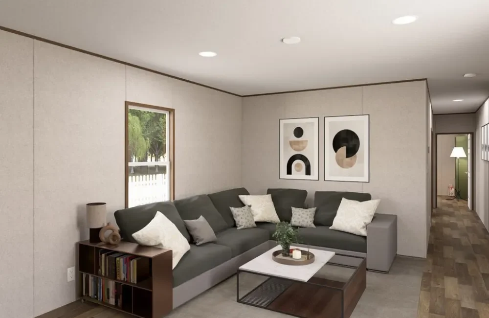 Living room with a gray sectional sofa with white and gray throw pillows, a small wooden side table with books and decorative objects, abstract art on the wall, a coffee table with a plant and candles, and a window showing greenery outside.