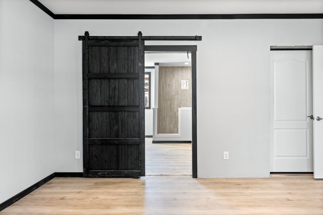 Interior of a room with a black sliding barn door, white walls, and a partially visible white interior door on the right. Light wood flooring throughout.