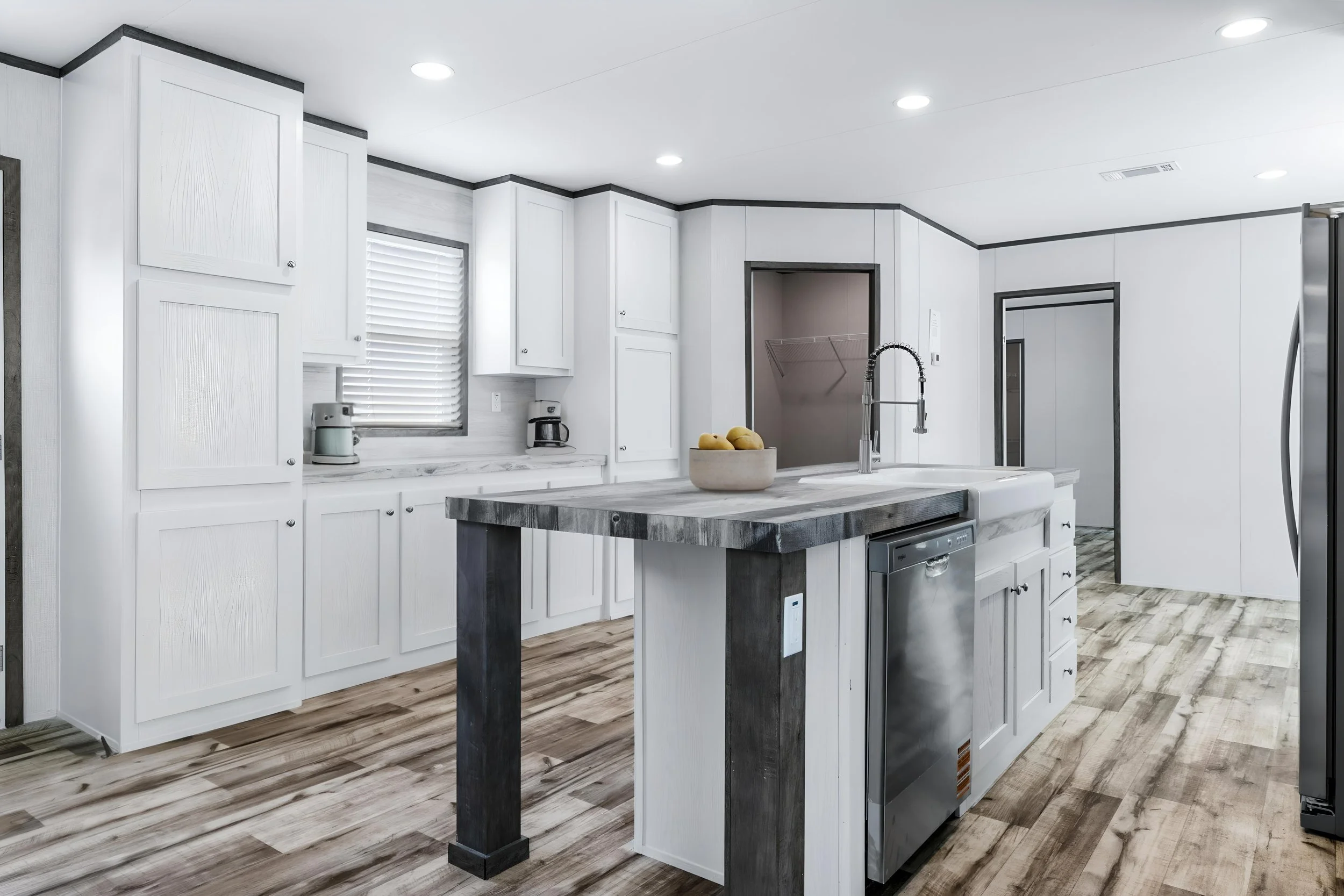 Modern kitchen with white cabinets, a kitchen island with a gray top, a stainless steel dishwasher, a coffee maker, a bowl with yellow lemons, a window with blinds, and wood flooring.