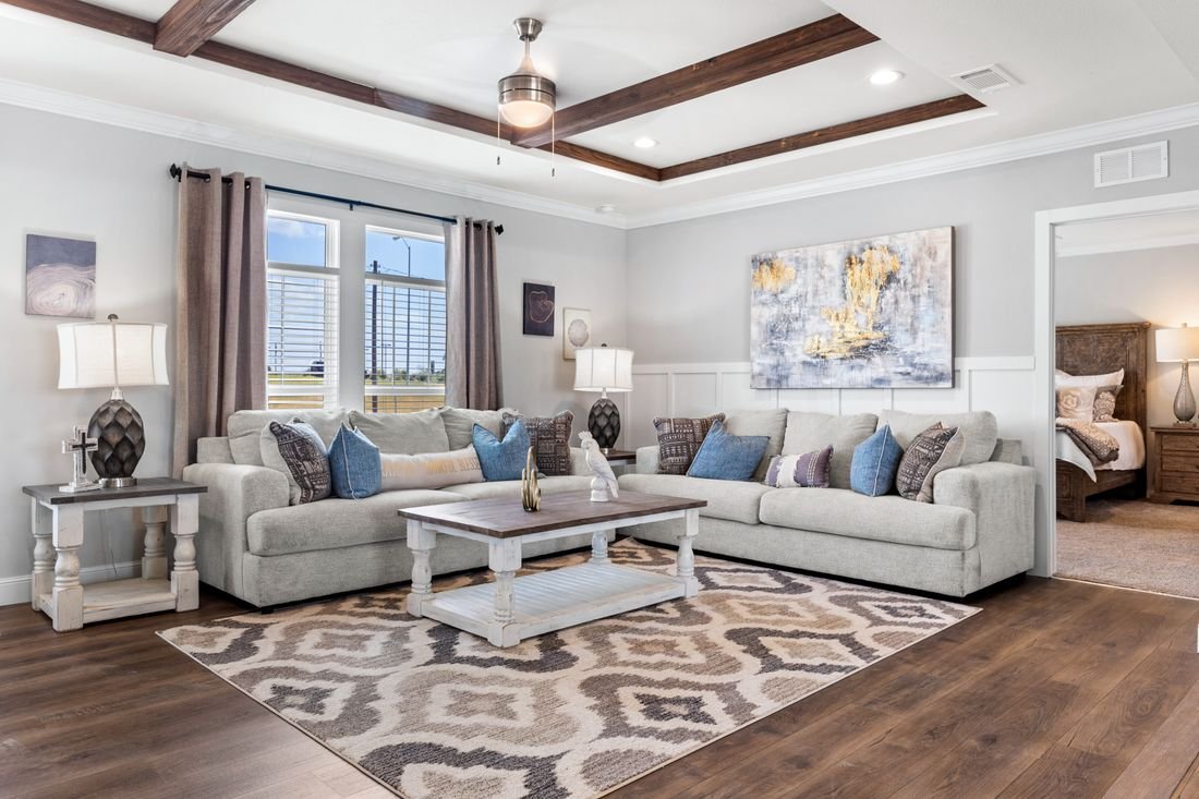 Living room with light-colored sofas, throw pillows, two side tables with lamps, a patterned area rug, a coffee table, large window, and a bedroom visible through an open doorway.