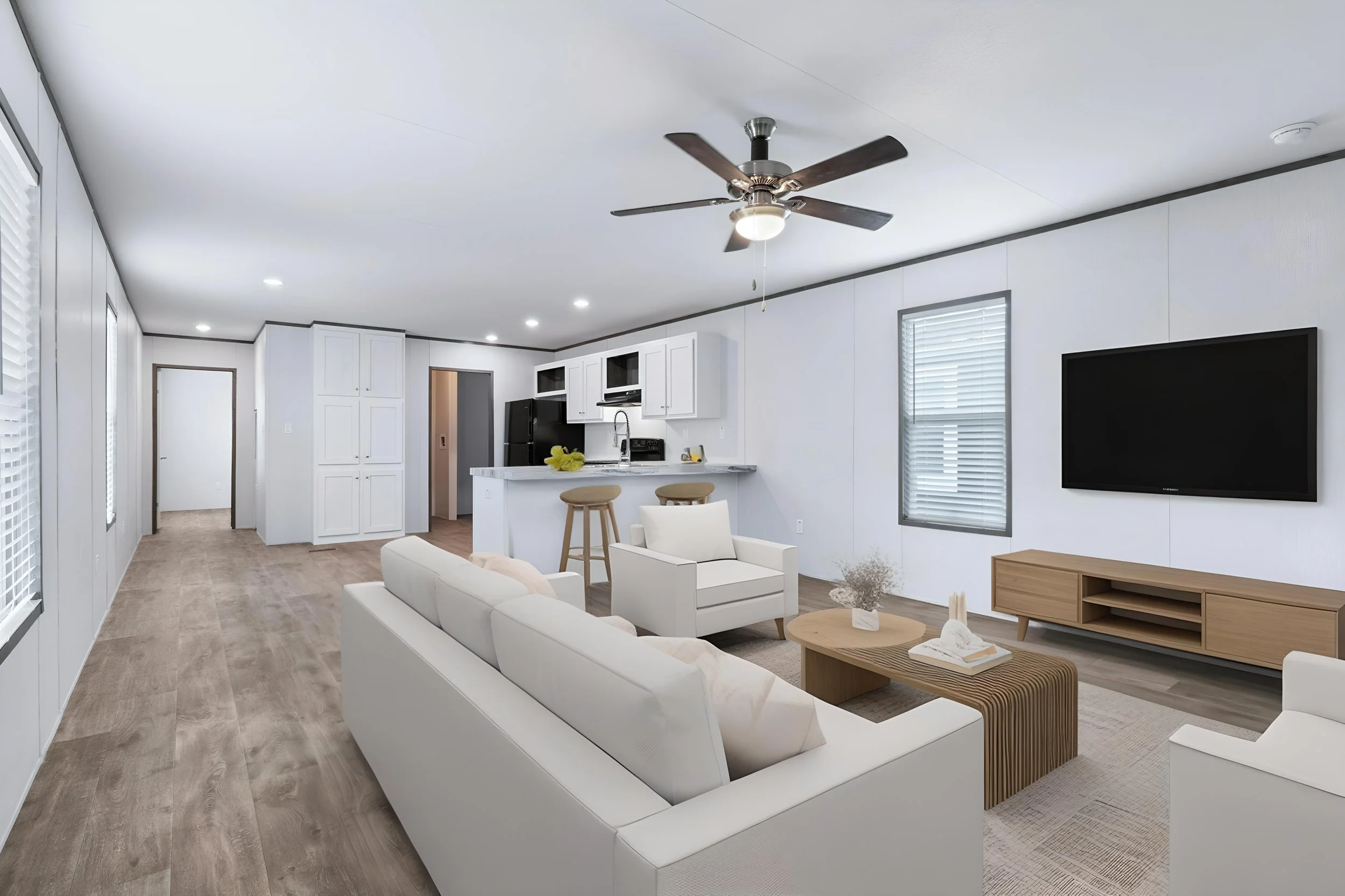 Living room with white sofas, a wooden coffee table, a large flat-screen TV, windows with blinds, a ceiling fan, and an open kitchen in the background.