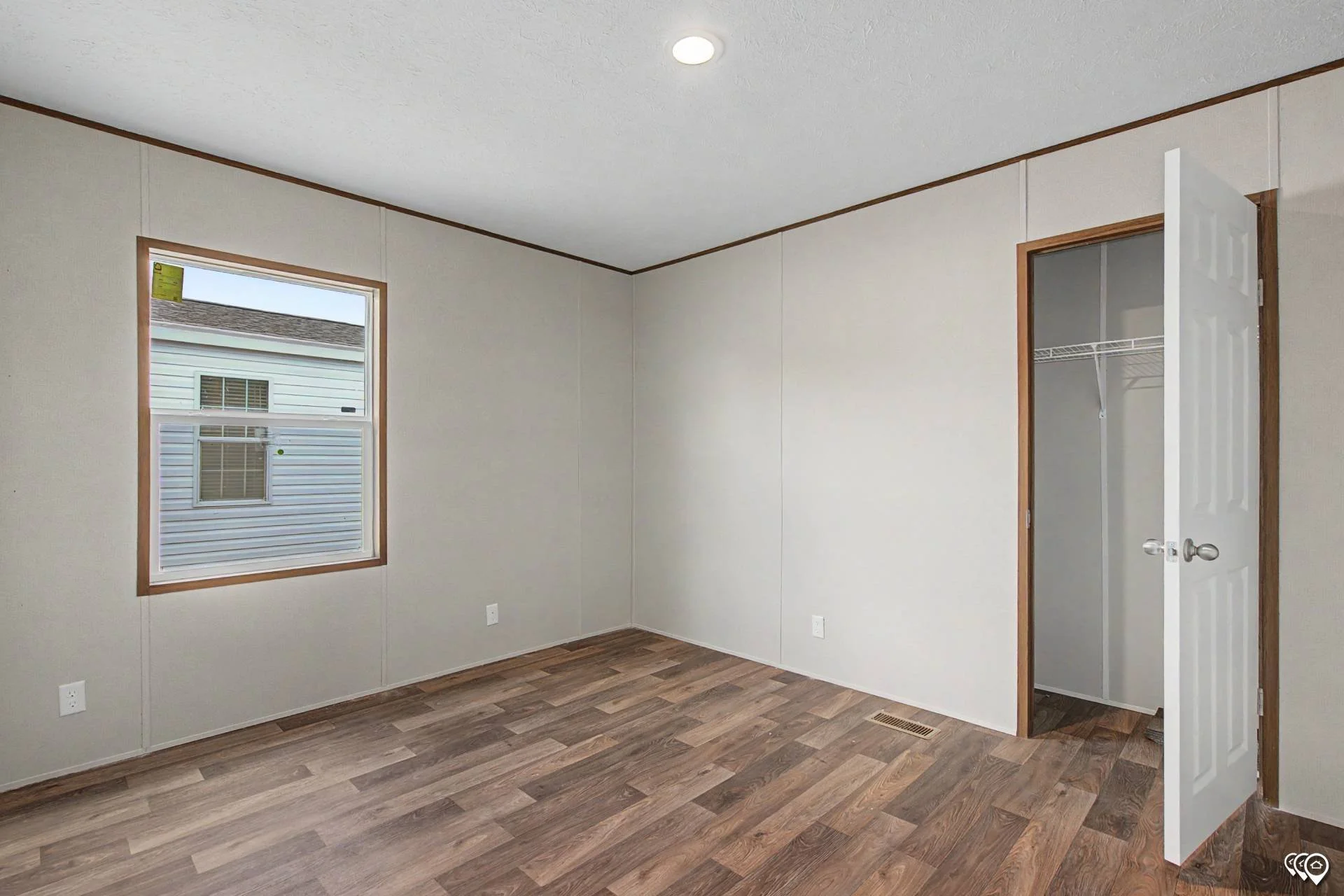 Empty room with beige walls and brown wood flooring, a window on the left wall, and an open closet door on the right wall.