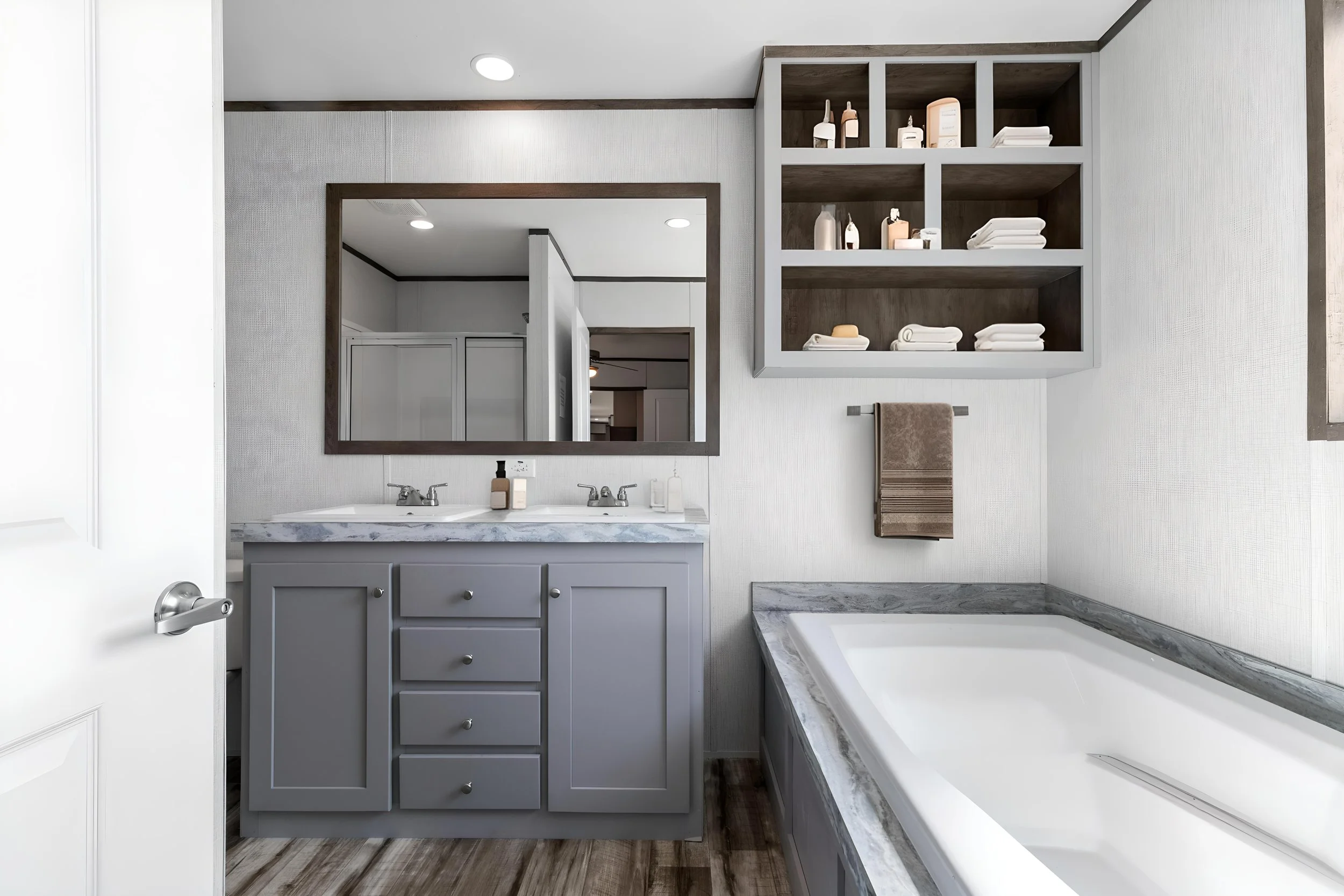 Bathroom with gray cabinet, marble countertop, large mirror, beige shelves with towels and decor, bathtub with marble surround, wood flooring, white walls, ceiling lights.