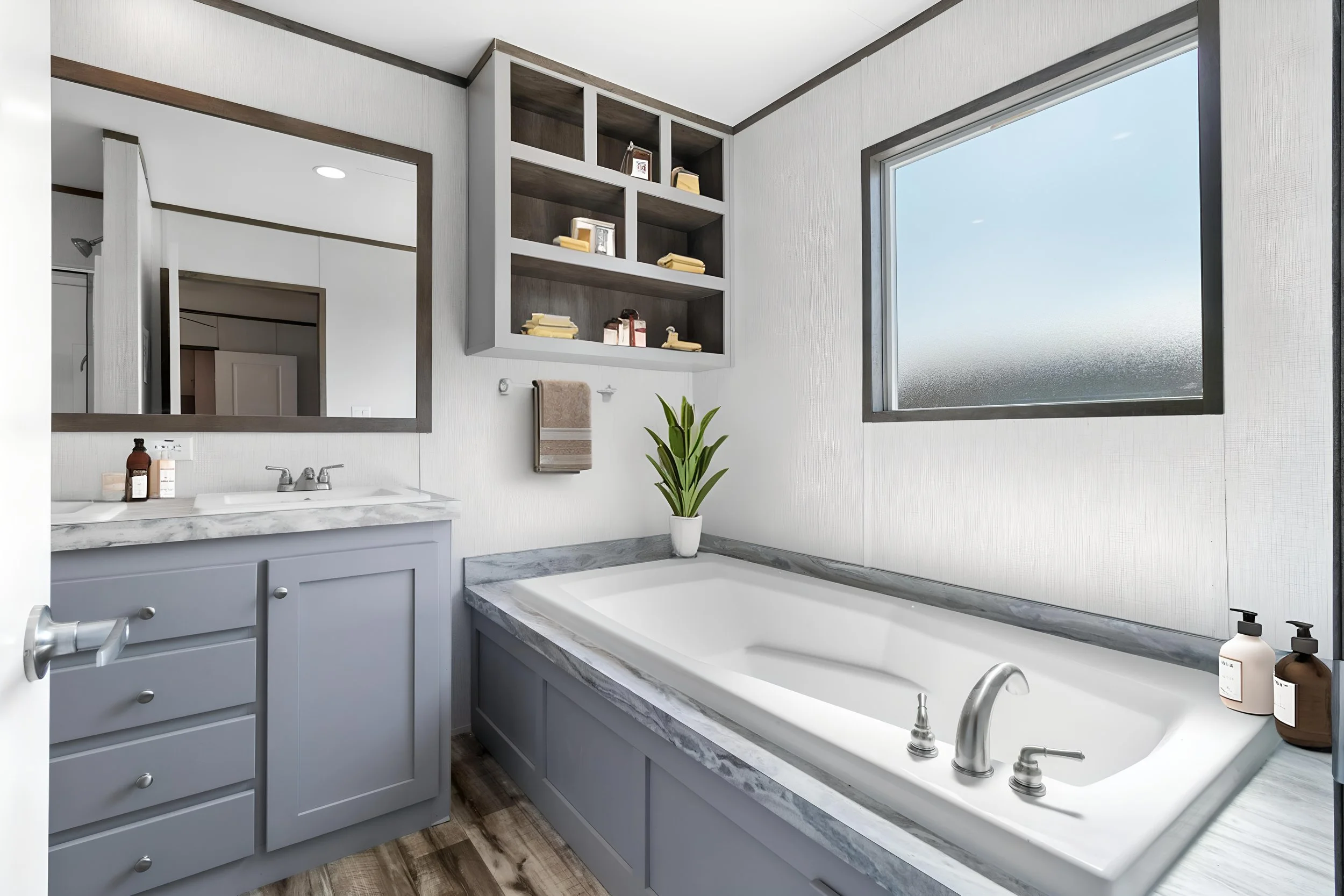 A modern bathroom featuring a large bathtub with a marble surround, a window with frosted glass, a vanity with a gray cabinet, a mirror, and open shelves with toiletries, a potted plant, and towels.