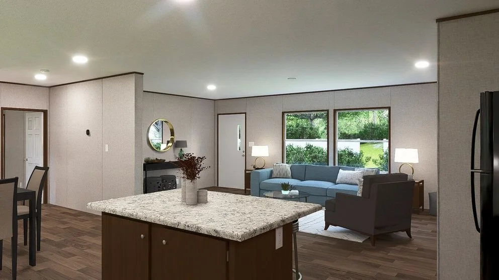 Open-concept living room and kitchen with a granite kitchen island, a blue sofa, a gray armchair, side tables with lamps, a round mirror on the wall, and large windows showing greenery outside.