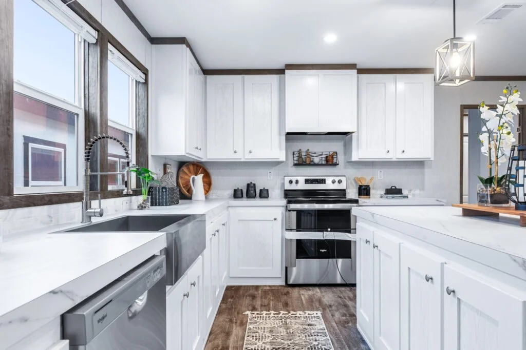 Modern white kitchen with stainless steel appliances and dark wood flooring, featuring a large window over the sink.