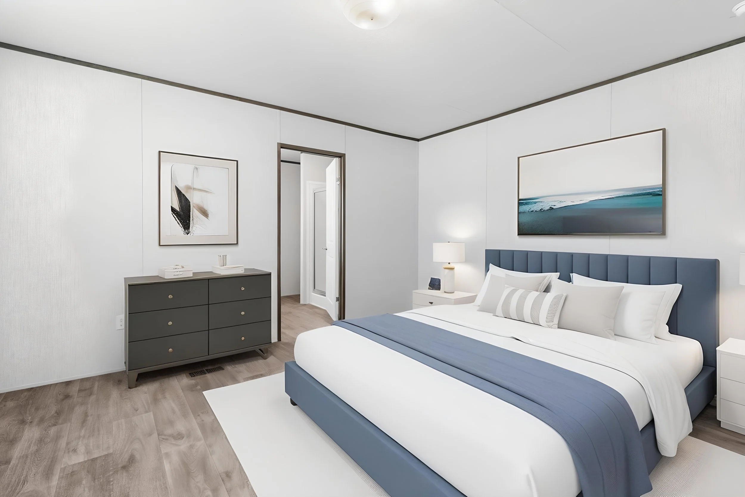 Modern bedroom with white walls, a bed with white and gray bedding, a navy blue upholstered headboard, a gray dresser with gold knobs, wall art, and a white nightstand with a lamp.
