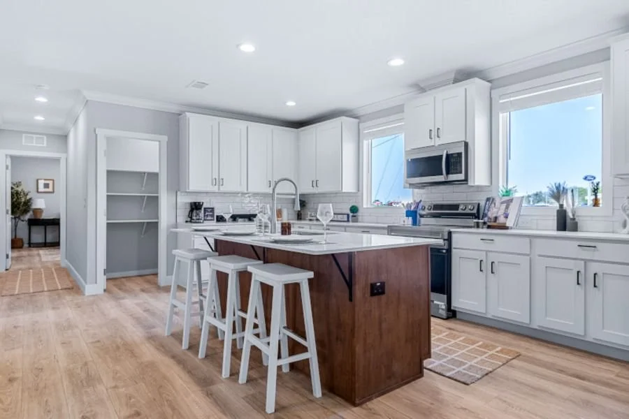 Bright, modern kitchen with white cabinets, a wood island with stools, stainless steel appliances, hardwood flooring, and large windows letting in natural light.