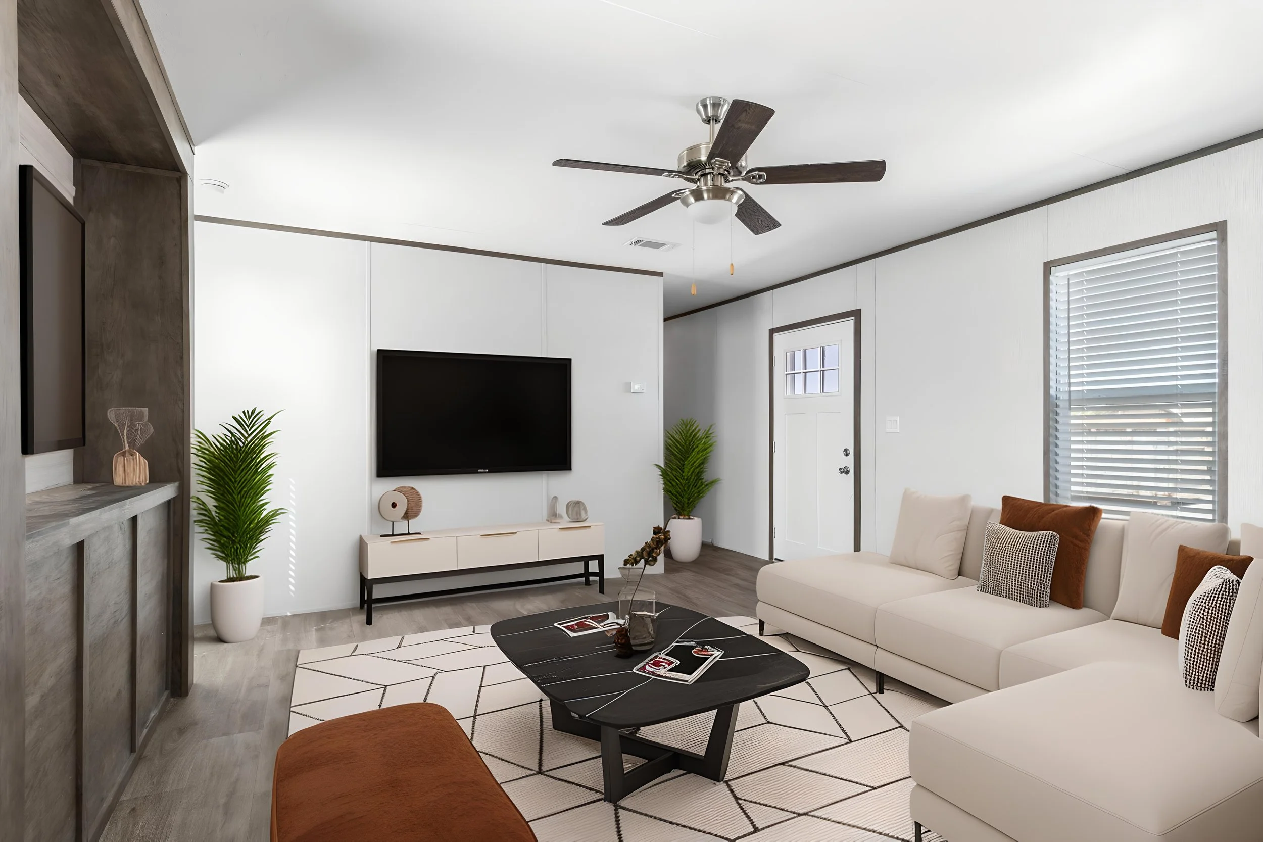 Living room with white walls and gray flooring, white sofa with patterned pillows, black coffee table, TV mounted on wall, potted plants, ceiling fan, window with blinds, door, and modern decor.