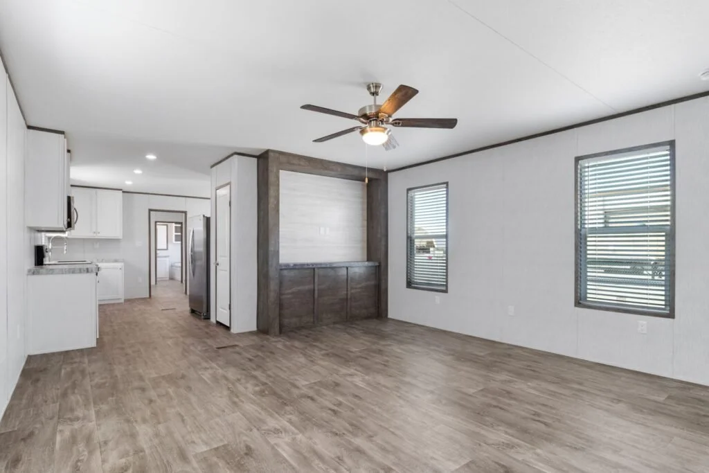 Empty living room with wooden floor, white walls, two windows with blinds, ceiling fan, and open kitchen area in the background.