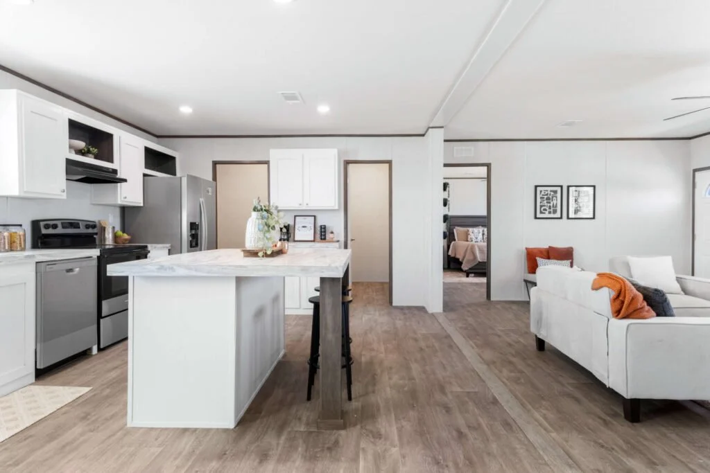 Open-concept kitchen and living room with white cabinets, stainless steel appliances, white sofa with orange and gray pillows, and wood flooring.