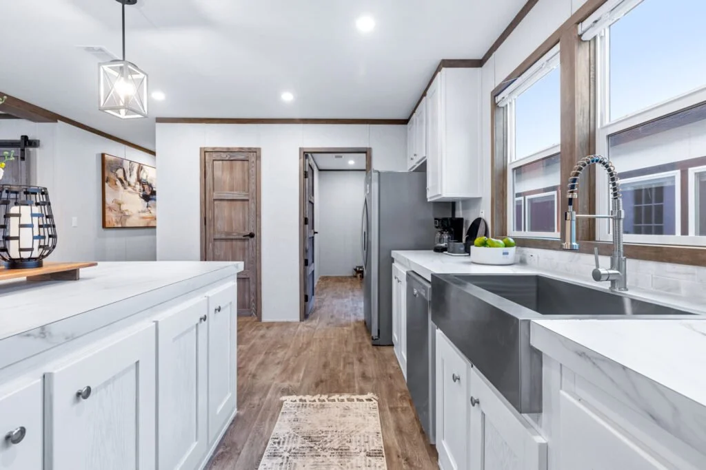 A modern kitchen with white cabinets, a large stainless steel sink under windows, a gray refrigerator, and wooden accents.