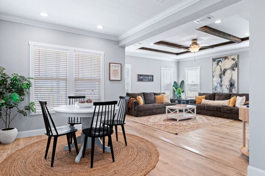 Bright living room and dining area with white walls, wooden floors, a round table with six black chairs, a brown sectional sofa, and various artwork and plant decor.