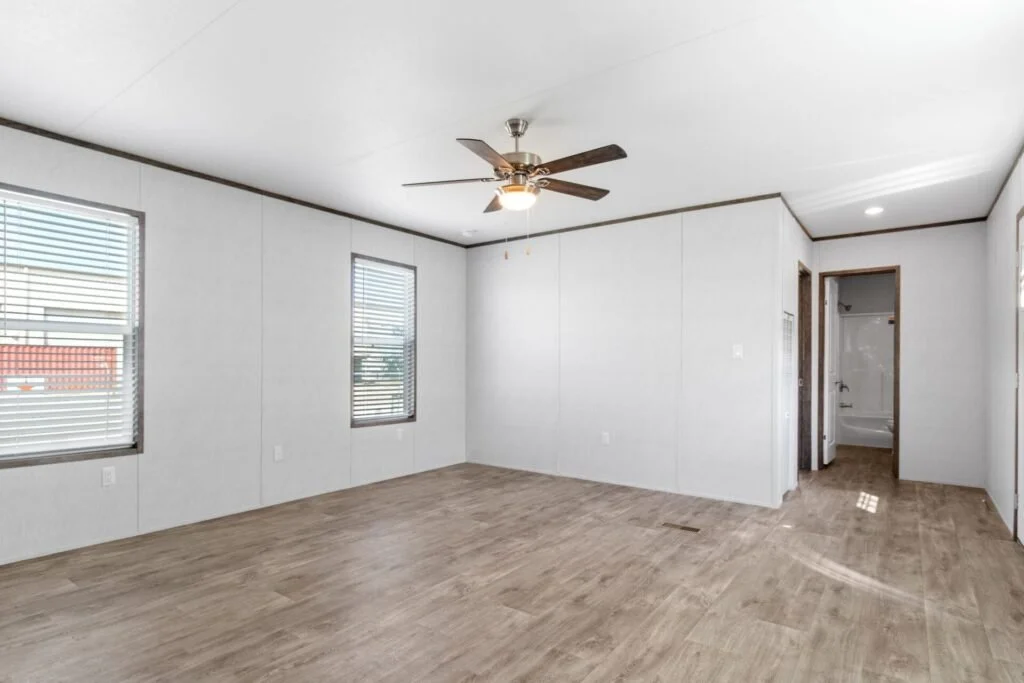 Empty living room with wood flooring, white walls, two windows with blinds, a ceiling fan, and an adjacent bathroom.