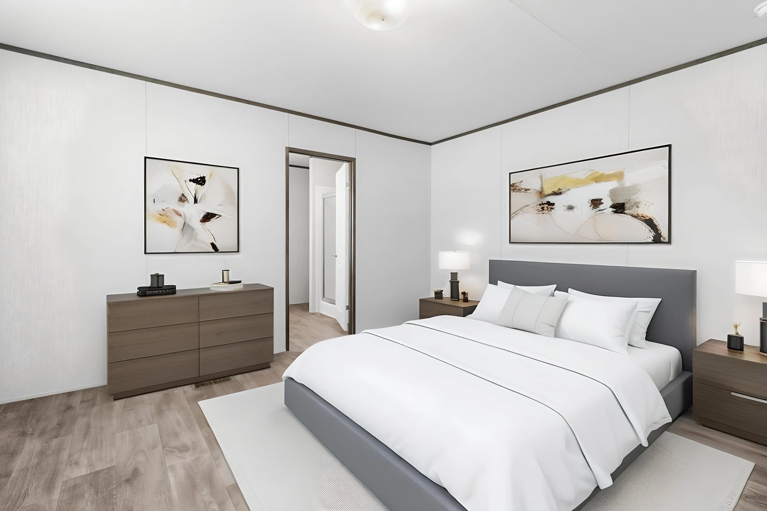 Modern bedroom with white walls, a gray bed with white bedding, two wooden nightstands with lamps, framed abstract artwork on the walls, and a doorway leading to another room.