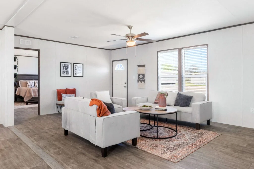 Living room with white sofas, a coffee table, and window blinds, leading to a bedroom with a bed and pillows.