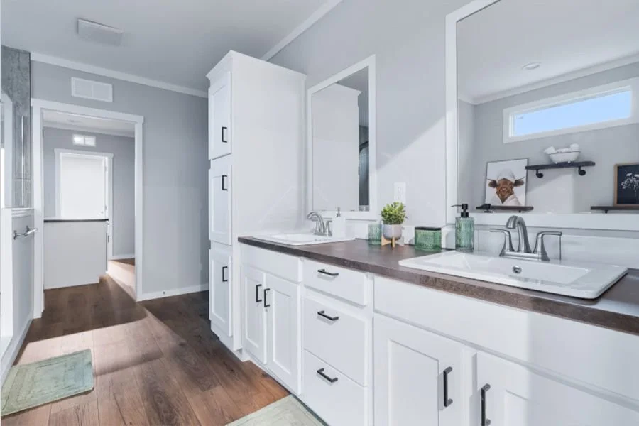 A bright, modern bathroom with white cabinets, dark countertop, and a large sink. Decor includes a small plant and soap dispensers. There is an open doorway leading to another room with wooden flooring and a window.