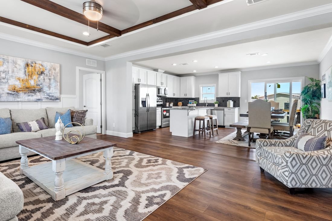 Open-concept living room and kitchen with hardwood floors, gray walls, white cabinets, stainless steel appliances, a dining table with four chairs, a beige armchair, a gray sofa with pillows, a large painting, a rug, and a ceiling fan.