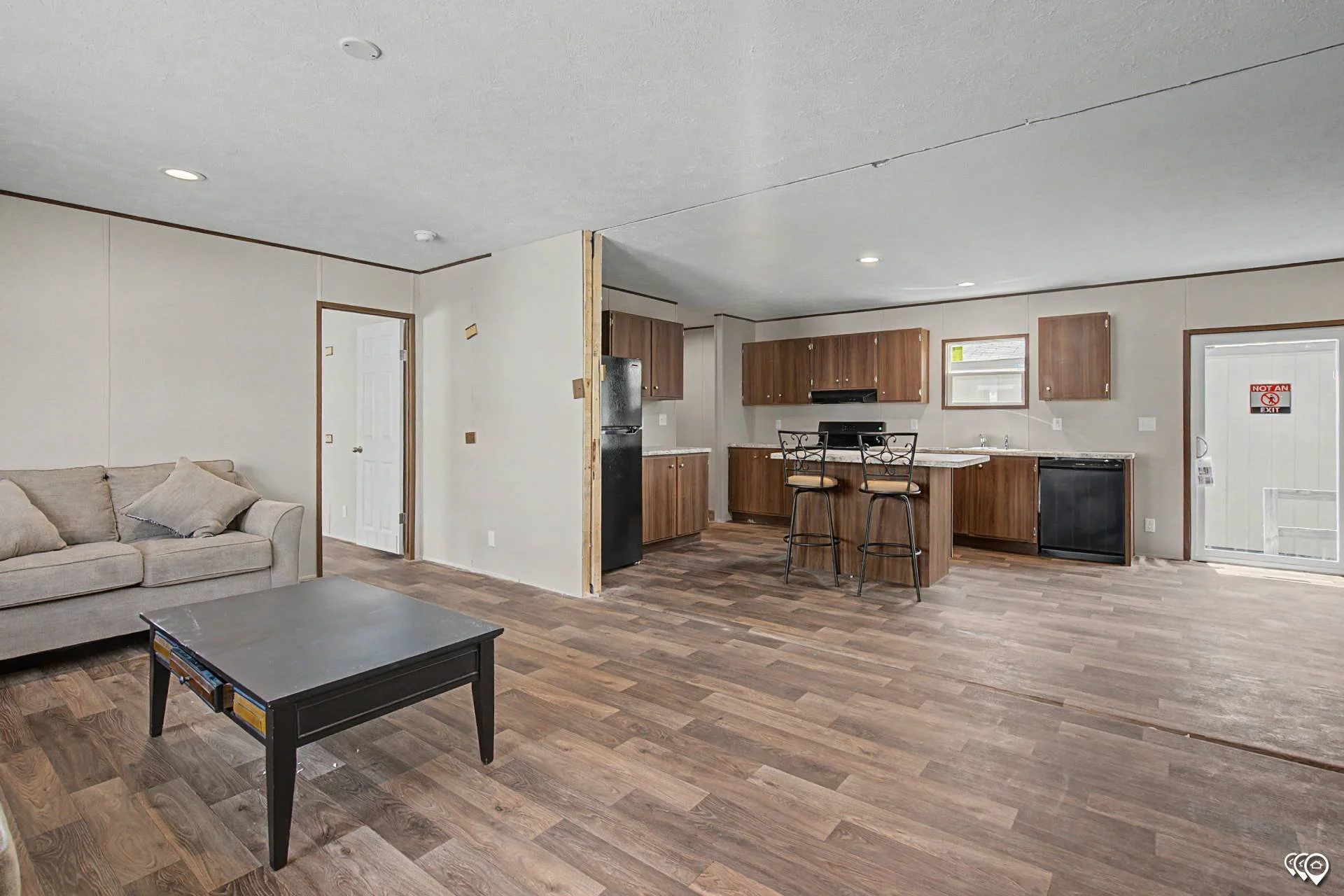 Open living area with brown wood flooring, beige sofa with pillows, black coffee table, and kitchen with wooden cabinets, black appliances, and barstools.