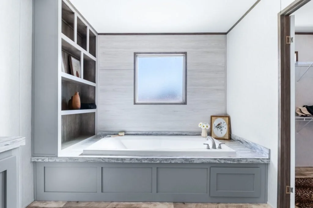 Bathroom with built-in bathtub, gray cabinetry, a window, and decorative items including framed pictures and a flower vase.