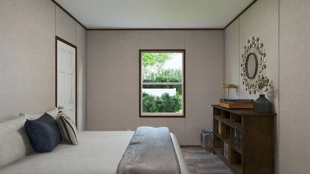 A bedroom with beige walls, a window with greenery outside, a bed with various pillows, a wooden bookcase, and a decorative mirror on the wall.