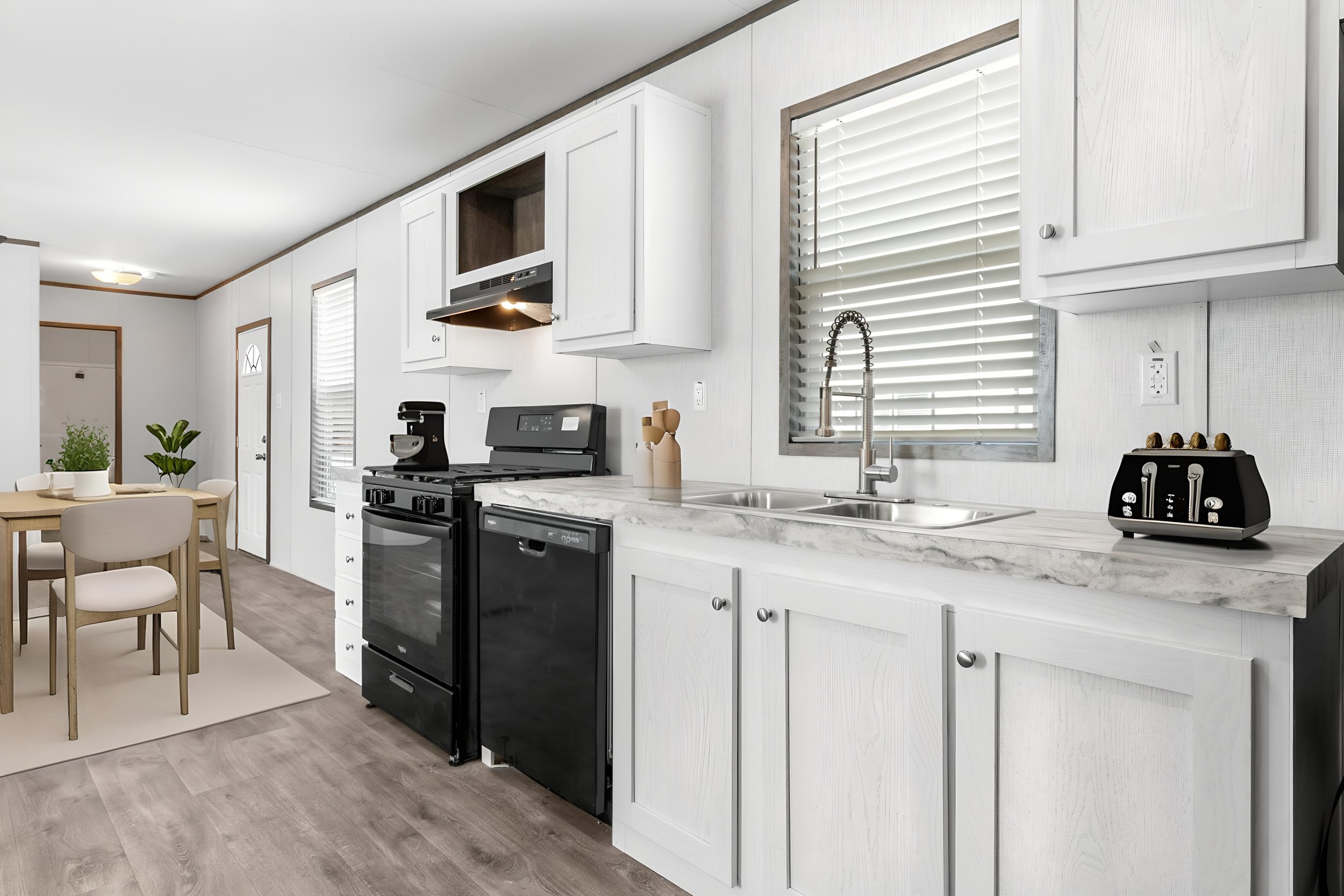 Modern kitchen with white cabinets, a black oven, a stainless steel sink, and a window with blinds. There is a small dining table with four chairs and some green plants.