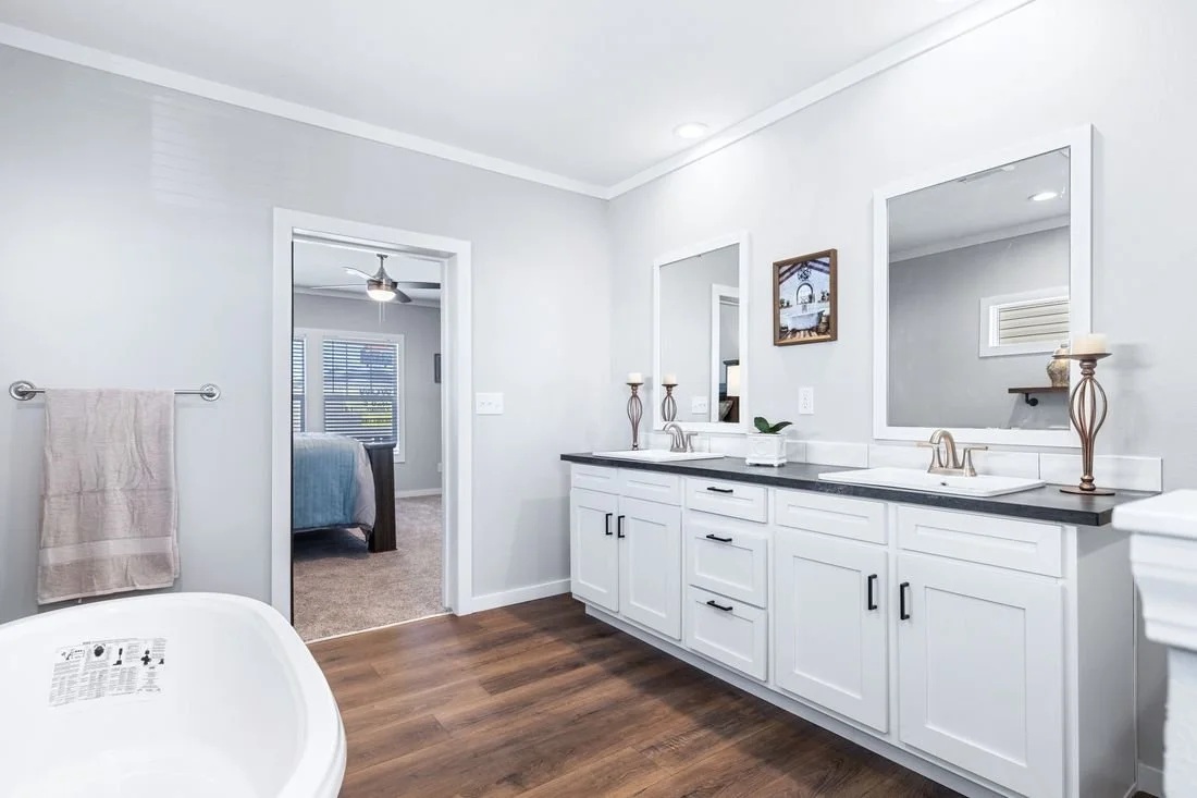 Modern bathroom with white cabinets, black countertops, large mirrors, and double sinks; includes candles and wall art, with a doorway leading to a bedroom.