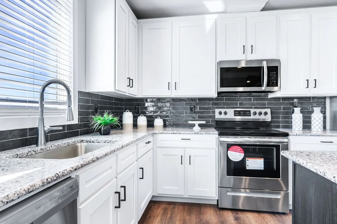 A modern kitchen with white cabinets, granite countertops, a stainless steel stove, microwave, and dishwasher, black subway tile backsplash, and a window with blinds.