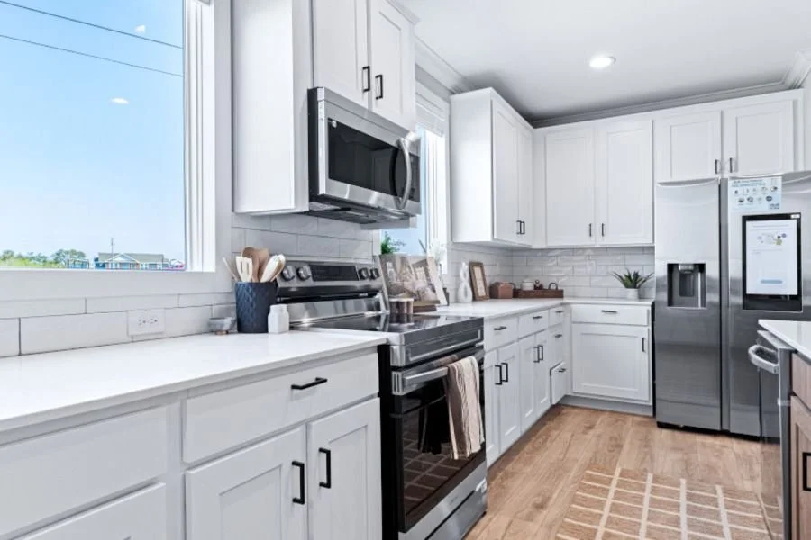 White kitchen with stainless steel appliances, including a microwave over the oven and a refrigerator, white cabinets, a window with a view outside, and a brown rug on wooden flooring.