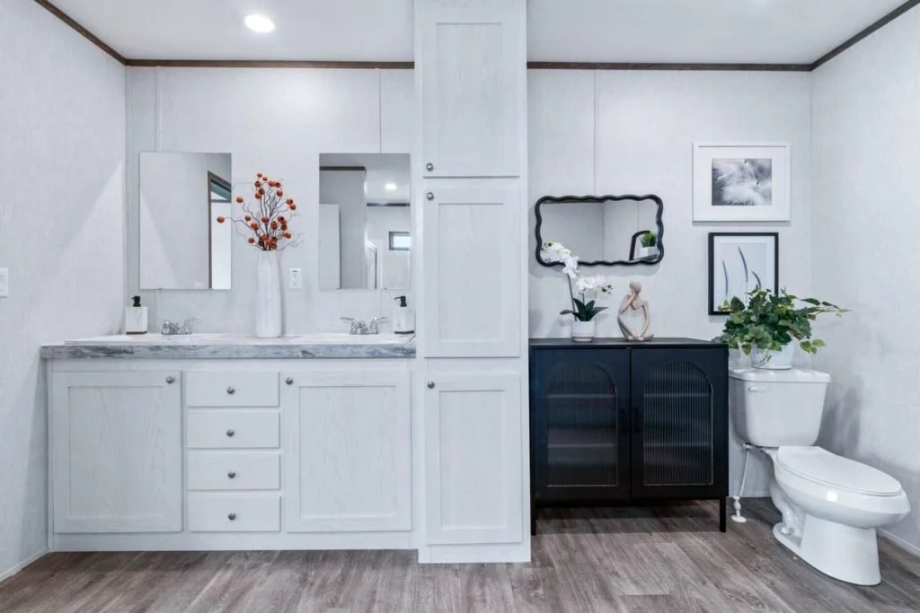 Modern bathroom with white vanity, marble countertop, dual sinks, wall mirror, black cabinet with decor plants, framed artwork, and a toilet, all on wood flooring.