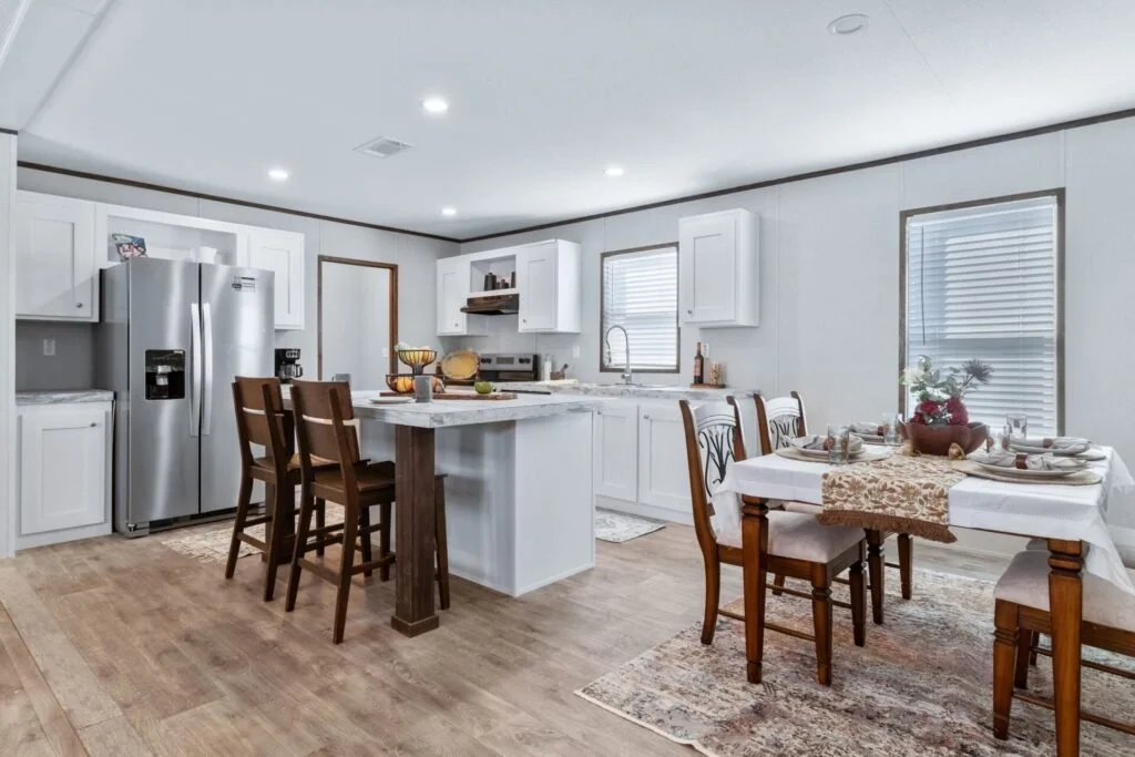 A dining area and kitchen in a home with white cabinetry, stainless steel refrigerator, and wooden furniture. The table is set with a tablecloth, decorative centerpiece, and place settings. The kitchen has a counter with fruit and a window with blind