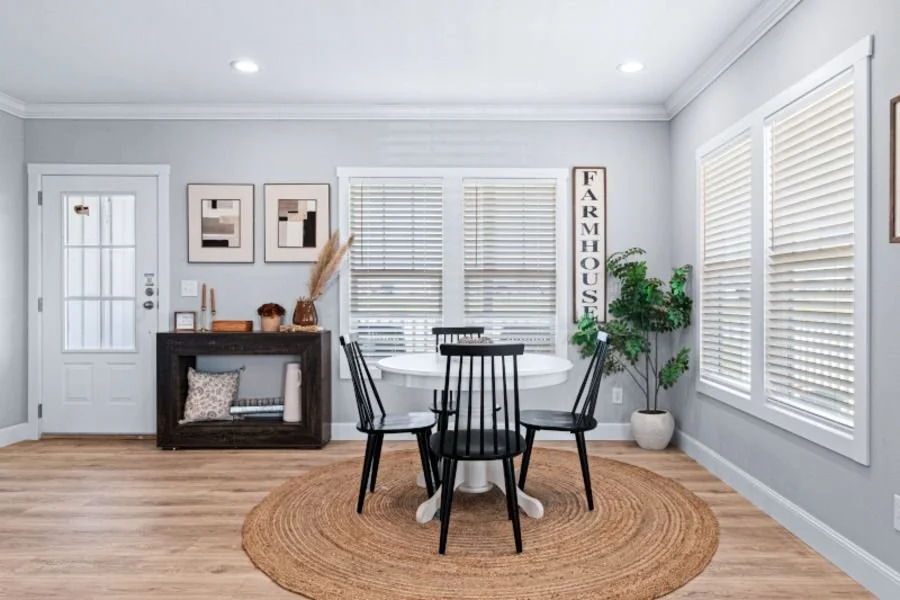 A cozy dining room with a round white table, four black chairs, a beige round rug, a black sideboard, framed artwork, a tall leafy plant, and a vertical sign that reads 'FARMHOUSE' in black letters on a white background.