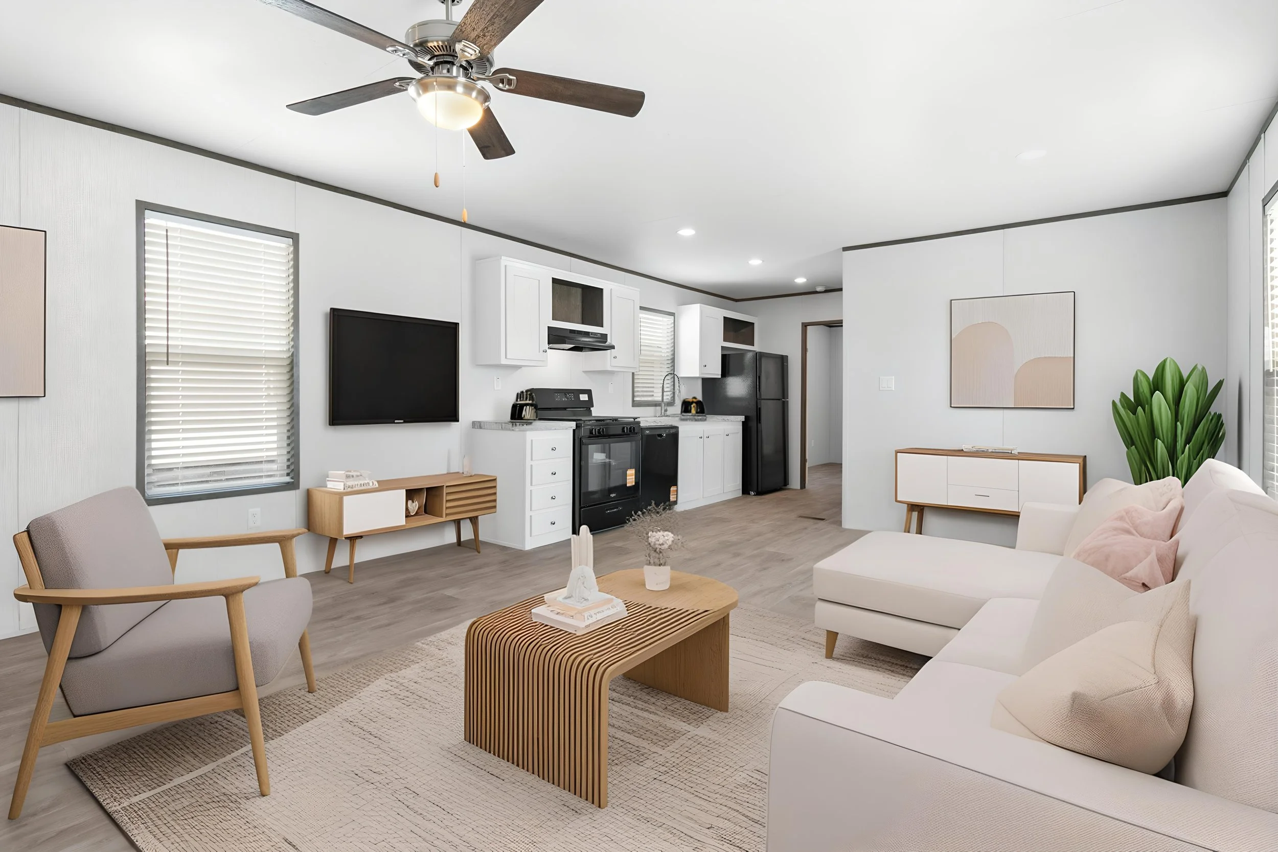 A modern, well-lit living room with white walls and ceiling, featuring a ceiling fan, a flat-screen TV on the wall, a kitchen with white cabinets and black appliances, a beige sofa with pink and beige pillows, a wooden coffee table, a gray armchair w