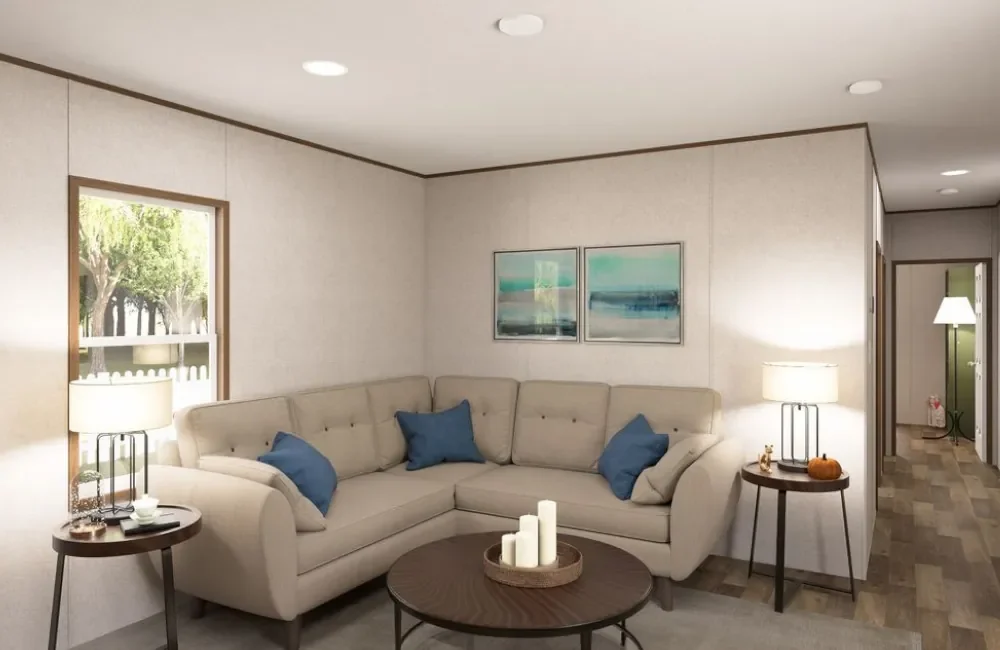 Living room with beige sectional sofa, blue pillows, two side tables with lamps, a coffee table with candles, window showing trees outside, and wall art with beach scenes.