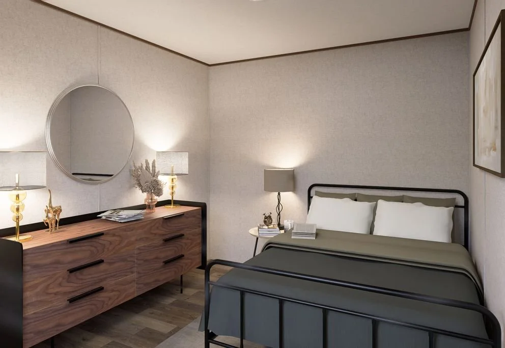A bedroom with a black metal bed frame, gray bedspread, white pillows, and a bedside lamp. A wooden dresser with black handles, two gold lamps, a mirror, and decorative items like dried flowers and a gold sculpture.