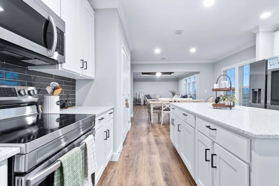 Modern kitchen with white cabinets, stainless steel appliances, gray subway tile backsplash, and a spacious open living and dining area in the background.