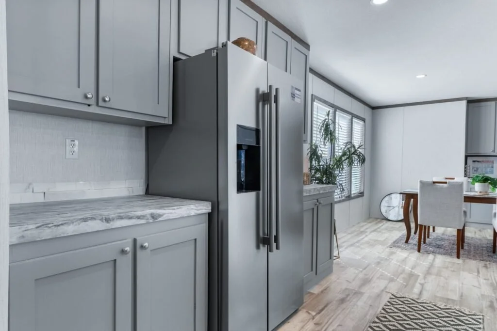 Modern kitchen with grey cabinets, stainless steel refrigerator, marble countertop, and dining table with chairs by windows.