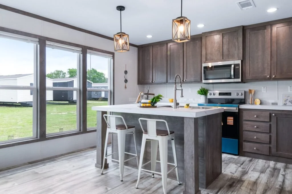 Modern kitchen with large windows, gray cabinets, a white island with two white chairs, pendant lighting, and stainless steel appliances.