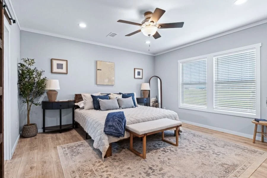 A bedroom with gray walls, a bed with beige and navy pillows, a bench at the foot of the bed, two windows with white blinds, a ceiling fan, and framed artwork on the walls.