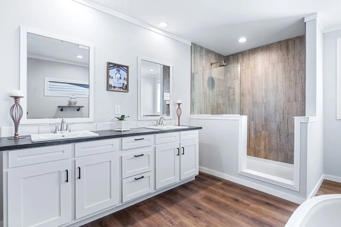 Bright bathroom with a white double vanity, black countertop, and two mirrors. There are decorative candle holders and a small plant on the vanity. A shower area with brown tiles and a glass enclosure is visible.