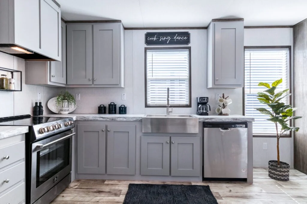 Modern kitchen with gray cabinets, a farmhouse sink, stainless steel dishwasher and stove, and decor including a sign that says "cook, snap, dance," a potted plant, and kitchen accessories.