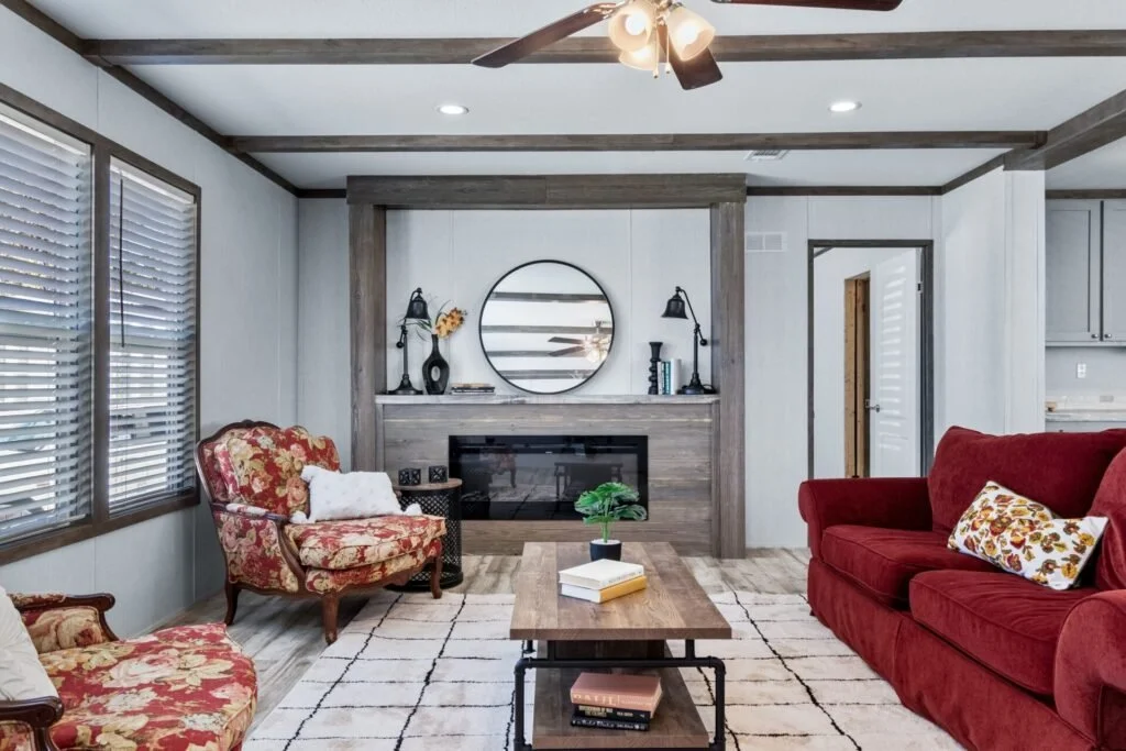 Living room with red and floral upholstered sofas, wooden coffee table, fireplace, round mirror on the wall, ceiling fan, windows with blinds, decorative items on the mantle, and a white rug.