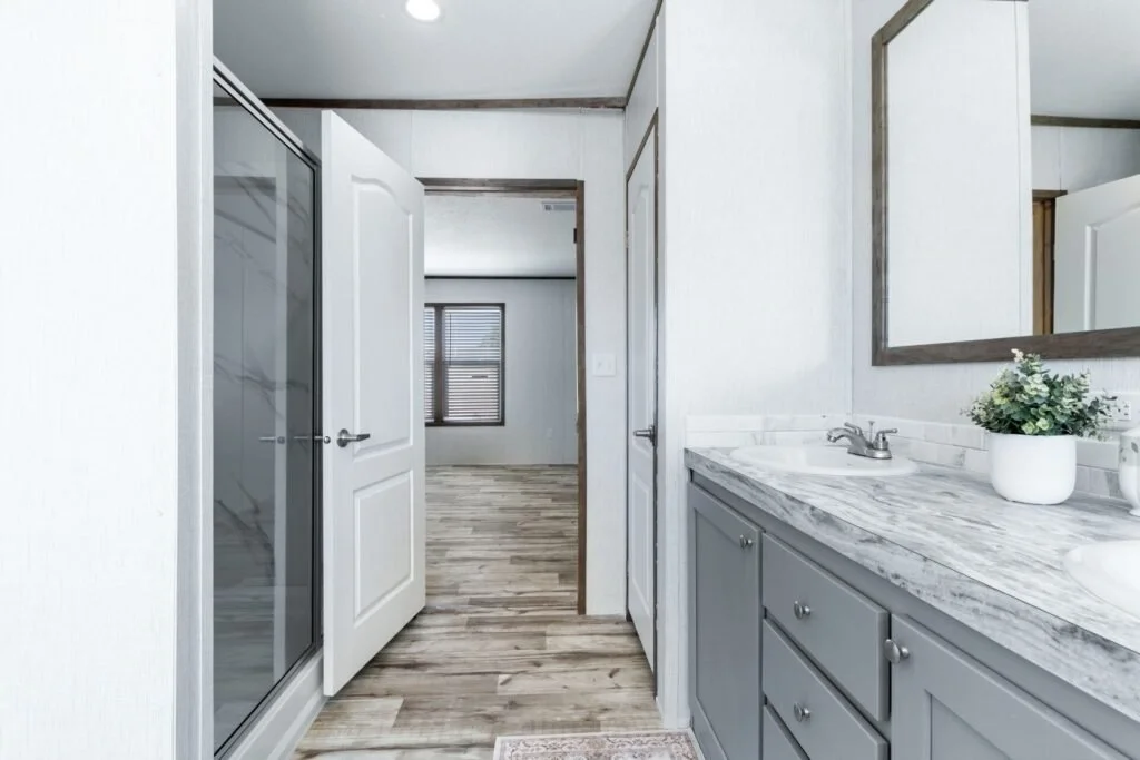 Bathroom vanity with a marble countertop, grey cabinets, a large mirror, and a small potted plant; open door leading to a room with a window
