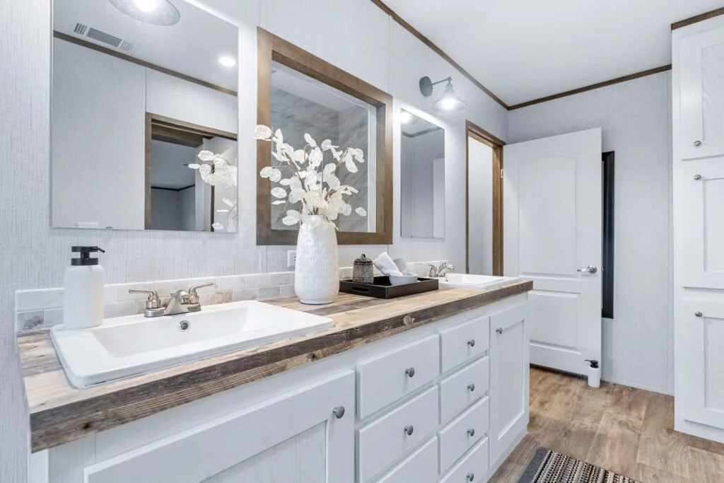A bright bathroom with a double vanity featuring a rustic wood countertop, white cabinets, two sinks, and large mirrors. Decor includes a vase with white flowers and a tray with toiletries. The room has grey wallpaper, wooden accents, and wood floori